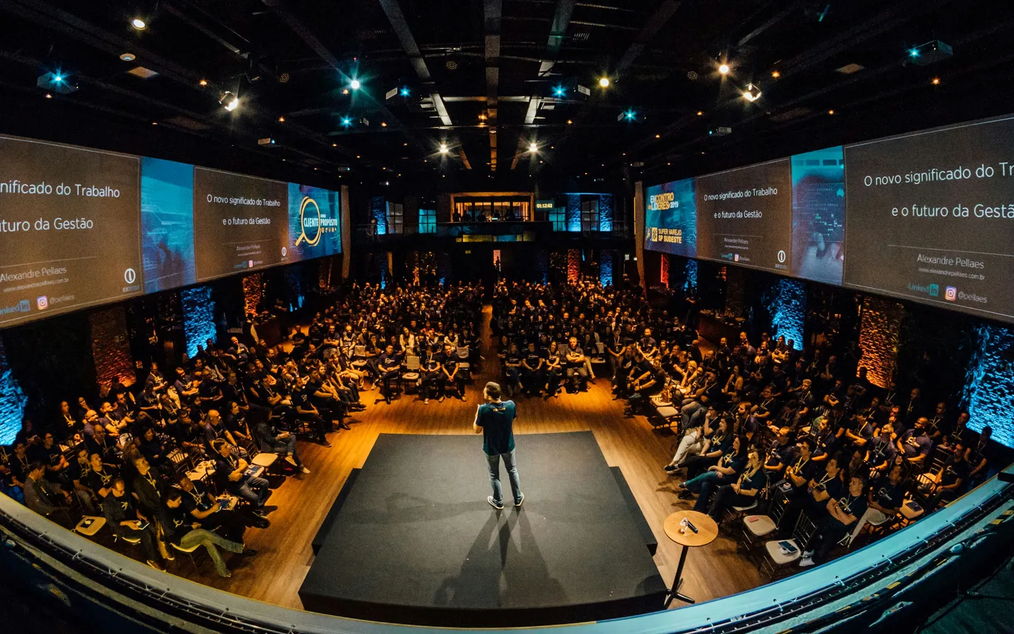 A speaker on stage addressing a packed conference hall in front of large presentation screens