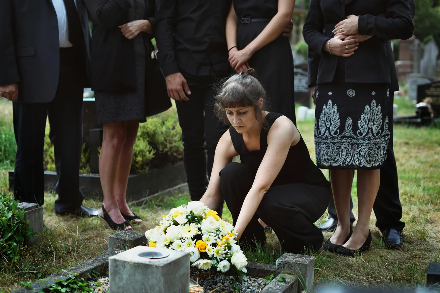 Mourners at a graveside as a woman kneels to lay a bouquet of white flowers
